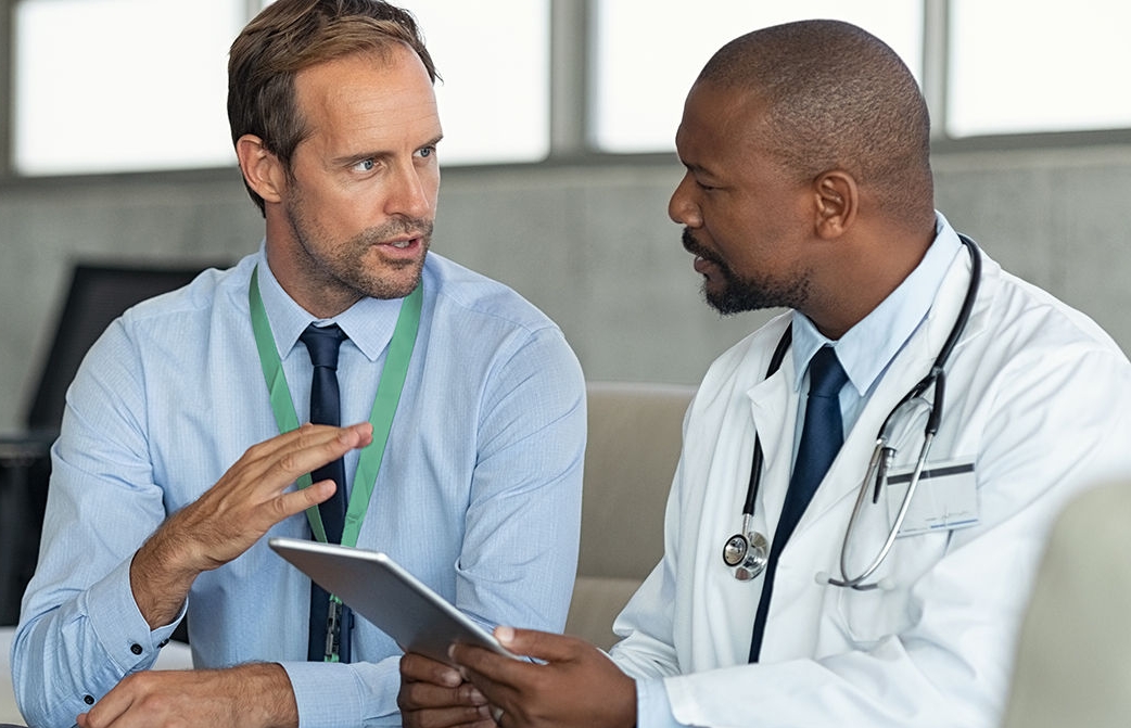 Pharmaceutical representative talking with doctor sitting on couch. African mature practitioner discussing results of the analysis with specialist while consulting diagnosis on digital tablet. Doctor in conversation with medical advisor in hospital room discussing patients report case.