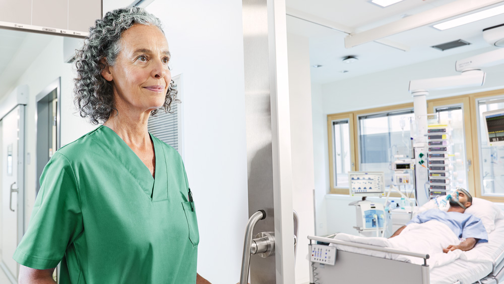 Person in medical scrubs stands outside an intensive care unit room with a patient in a hospital bed.